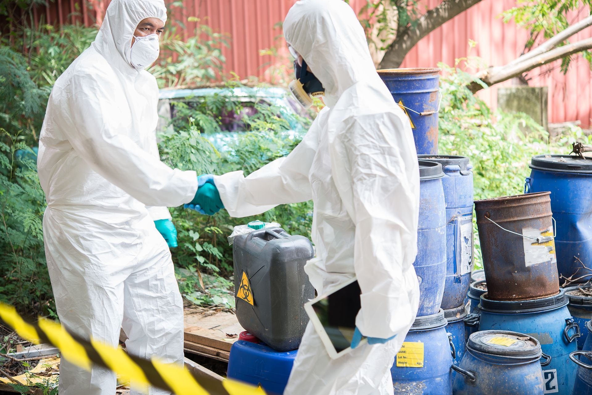 Two men in white hazardous material suits shaking hands in front of barrels labeled as hazardous waste.