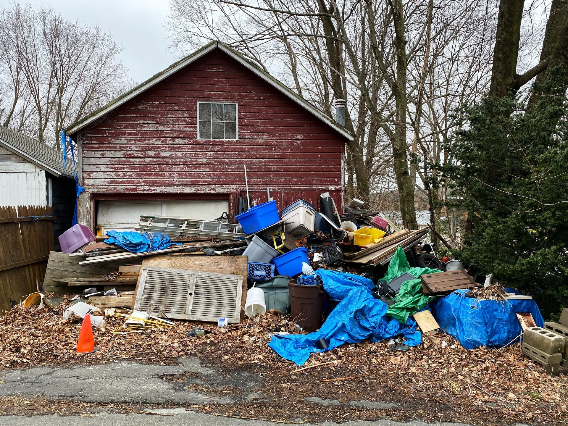 A red barn garage with a large, cluttered pile of debris, blue tarps, and trash scattered across its front yard.