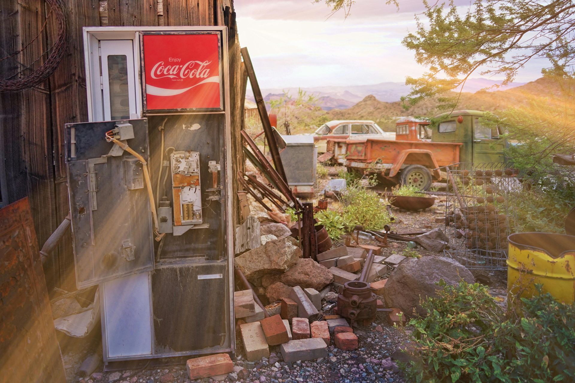 An abandoned Coca-Cola vending machine sits outdoors next to rusted scrap and a vintage orange truck at sunset.