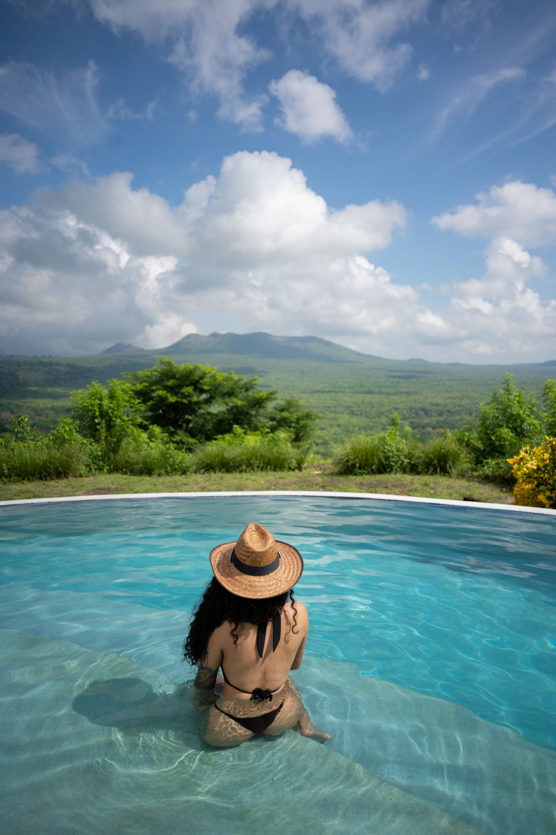 Person in a straw hat relaxing in a pool overlooking lush green mountains and a blue sky
