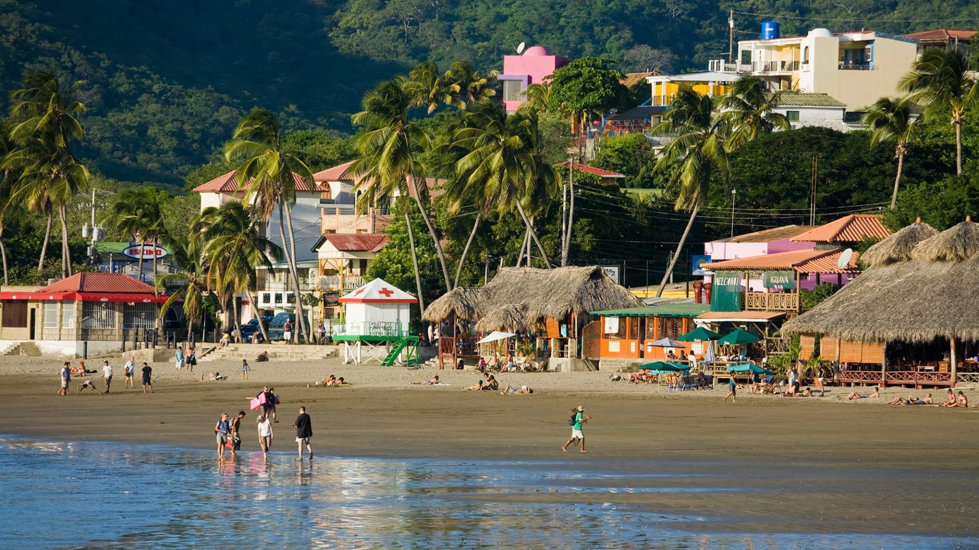 People on a sandy beach with palm trees, colorful buildings, and hills in the background