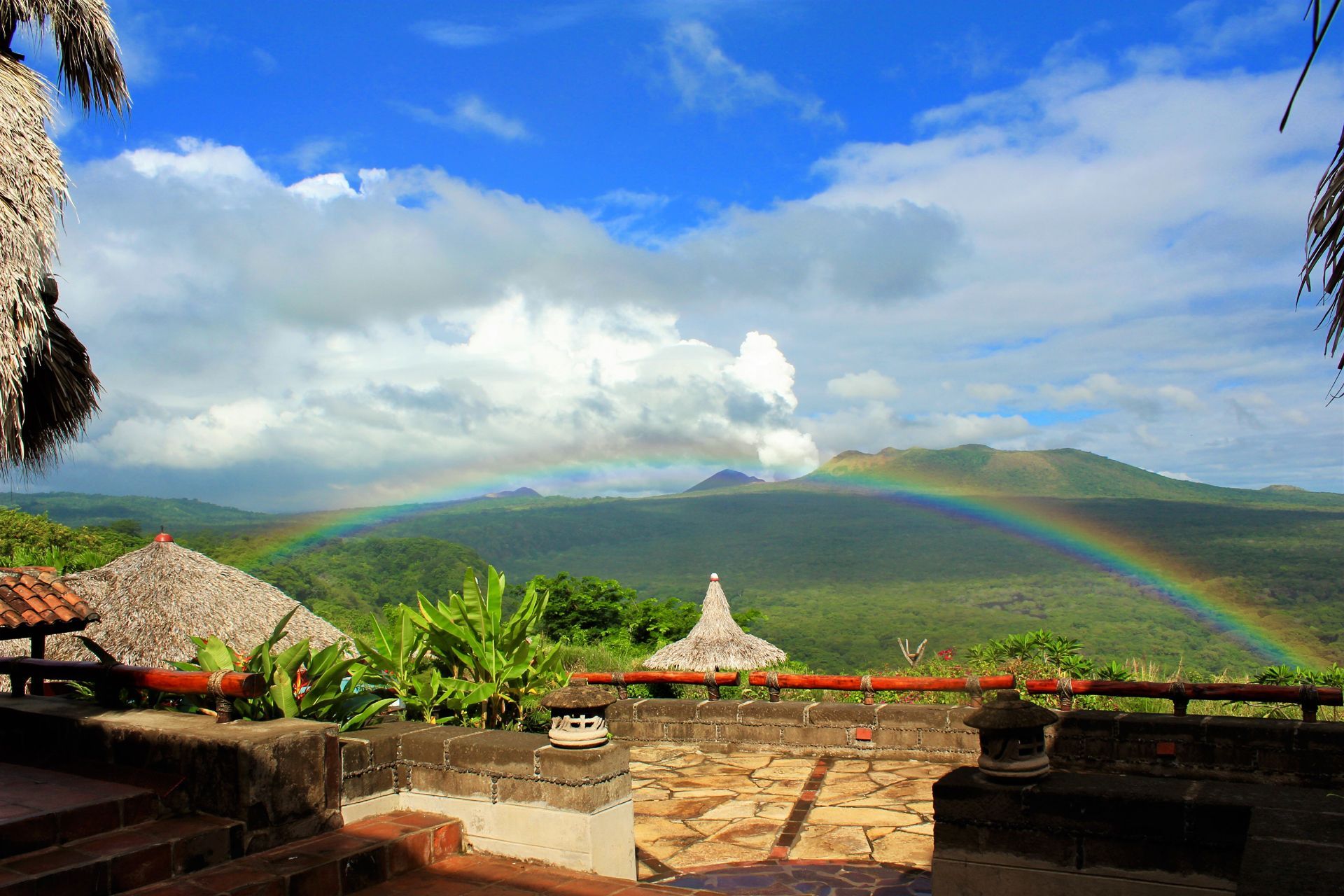 Tropical resort terrace with thatched huts, green mountains, blue sky, and a rainbow arching overhead