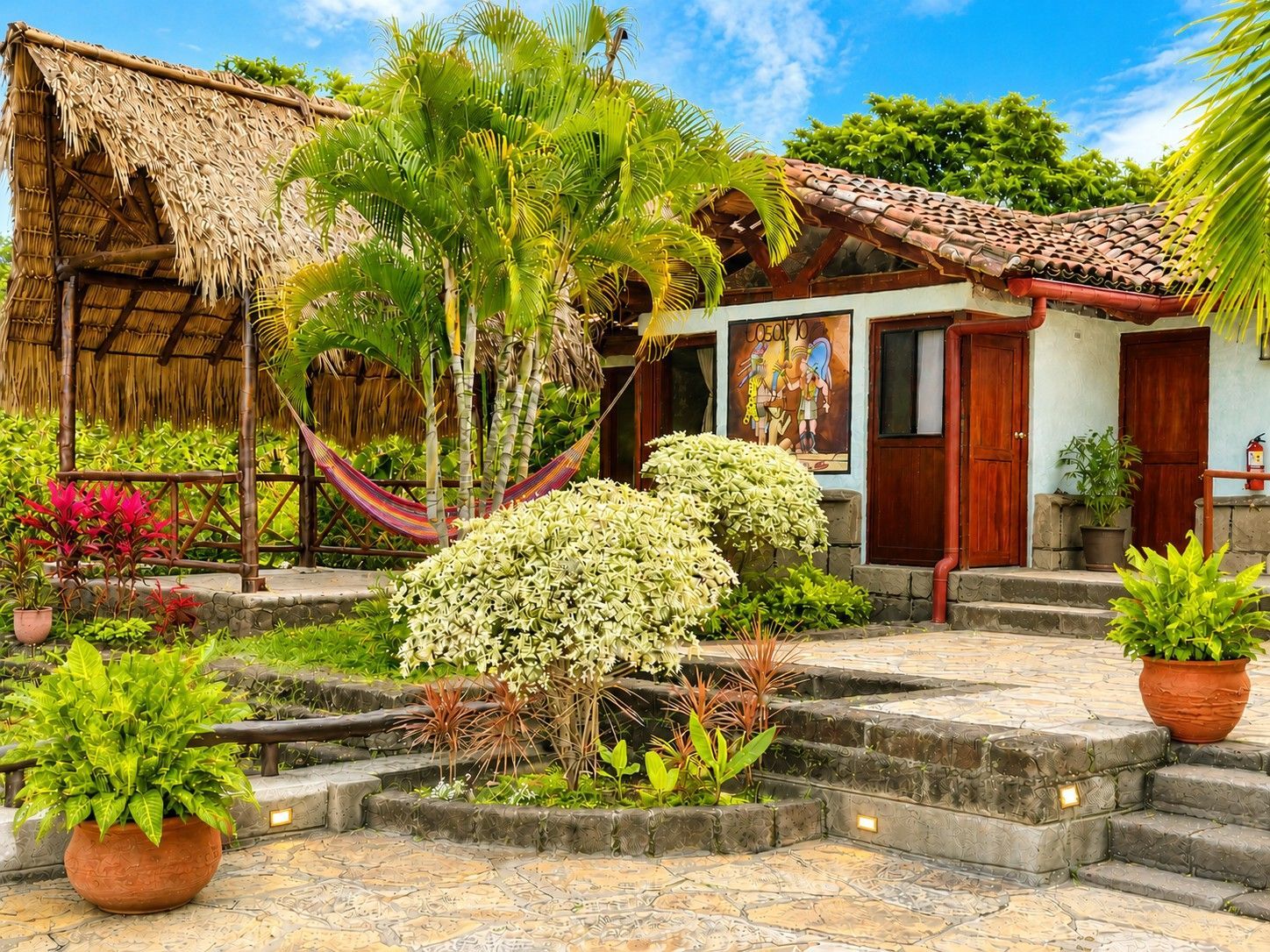 Tropical garden courtyard with thatched-roof cottage, palm trees, colorful flowers, and stone steps