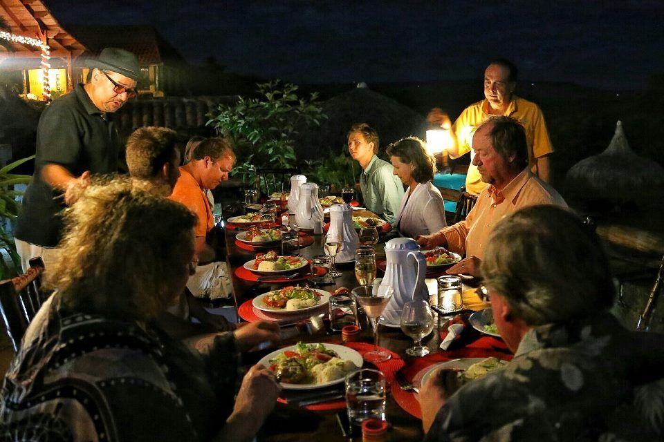 People dining outdoors at night under warm lights, seated around a long table with food and drinks.