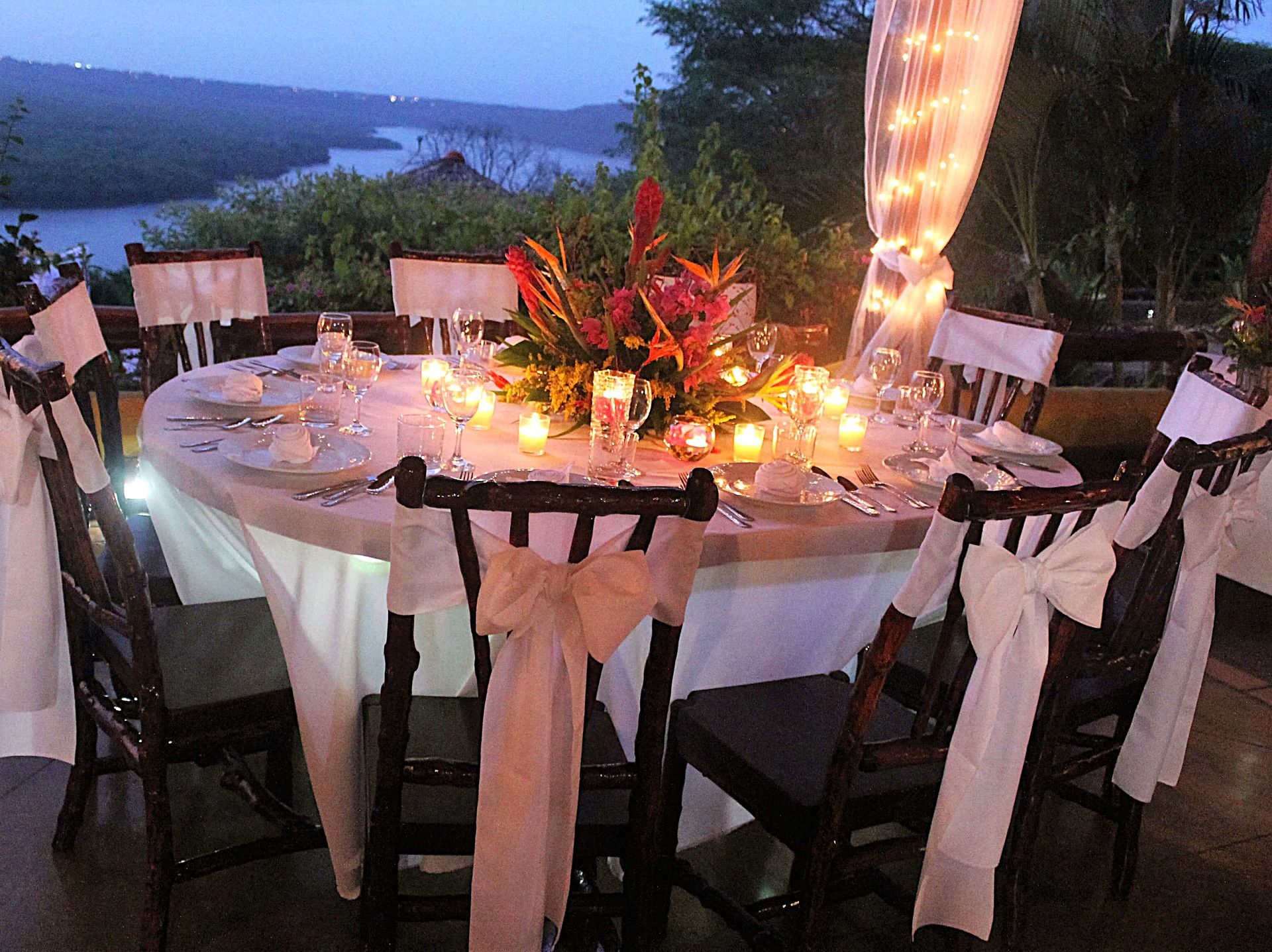 Lit dinner table outdoors at sunset with candles, flowers, and white-draped chairs by the water