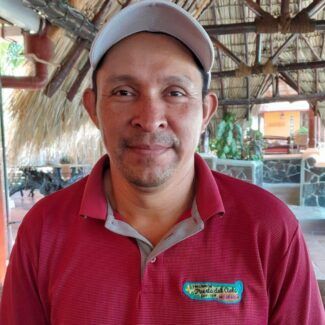 Man in a red polo shirt and cap smiling indoors at a rustic, open-air venue
