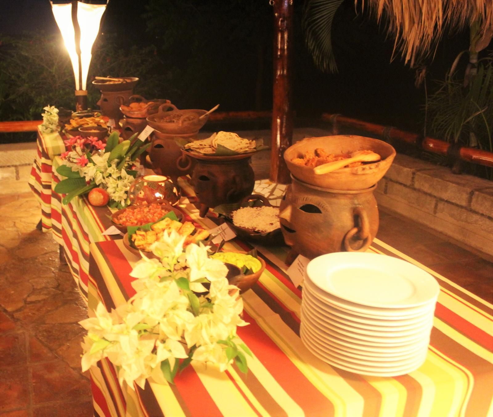 Buffet table with floral centerpiece, clay pots, and stacked white plates under warm lighting.