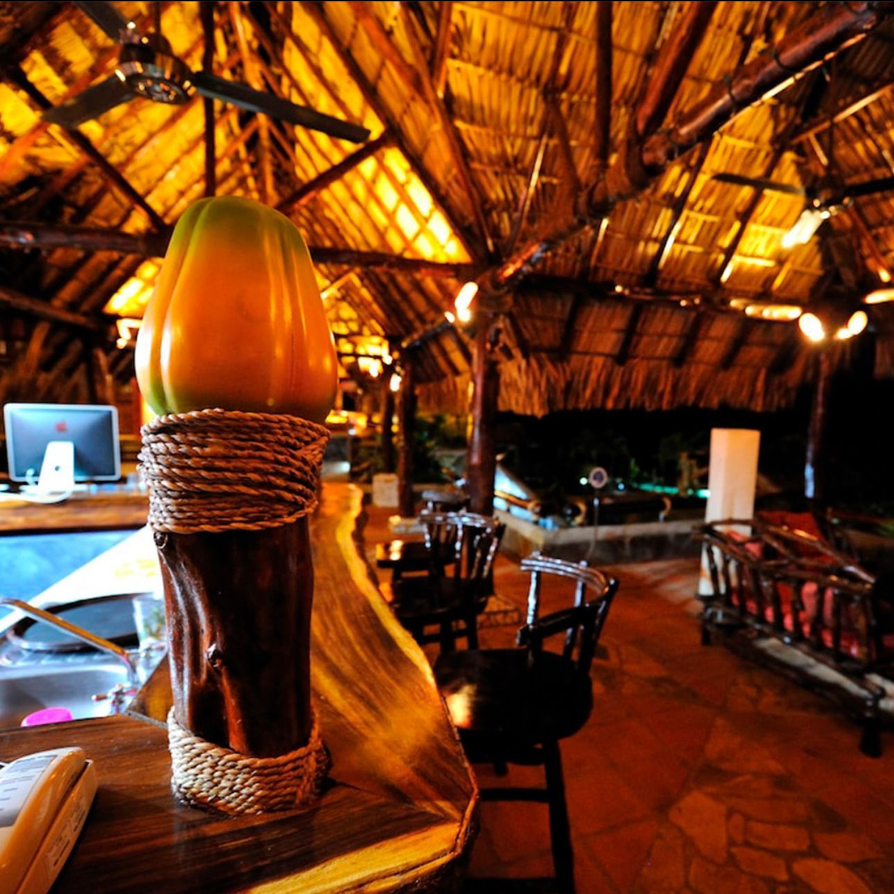 Warmly lit thatched-roof bar interior with wooden tables, chairs, and woven decor.