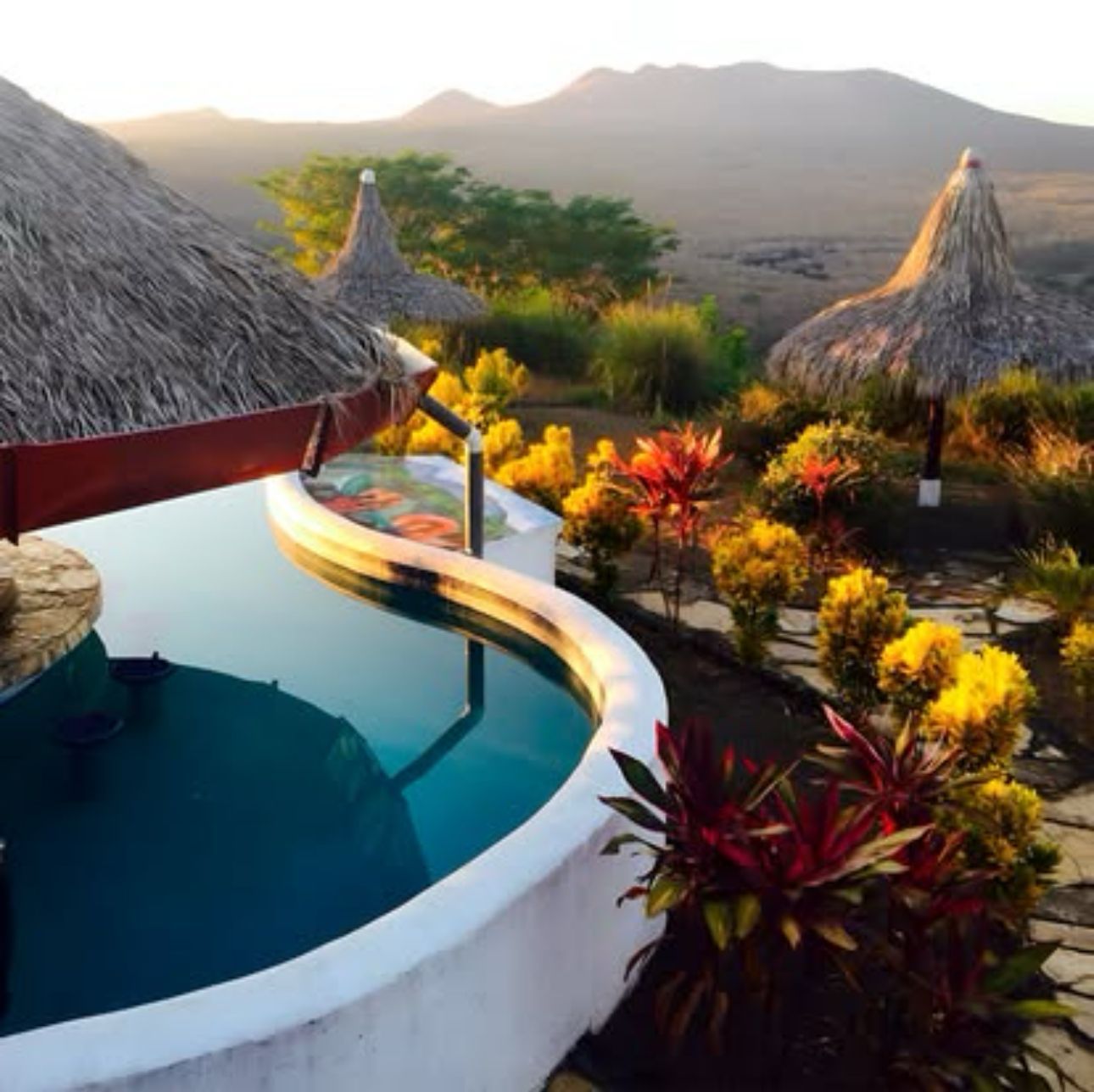 Curved tropical pool beside thatched huts and colorful plants at sunset, overlooking mountains