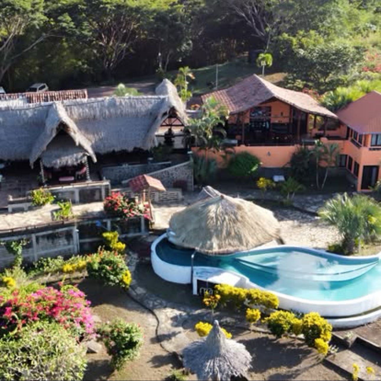 Tropical resort with thatched huts, a curving blue pool, and lush greenery on a hillside.