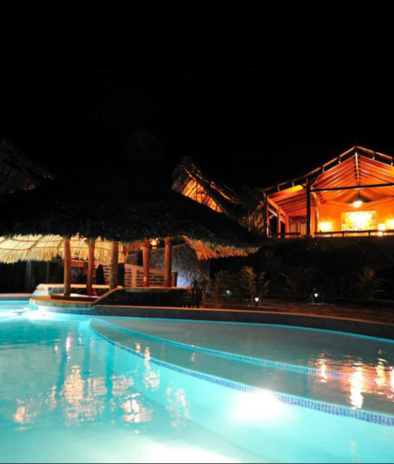 Nighttime resort pool with turquoise water and warmly lit thatched villas reflected on the surface