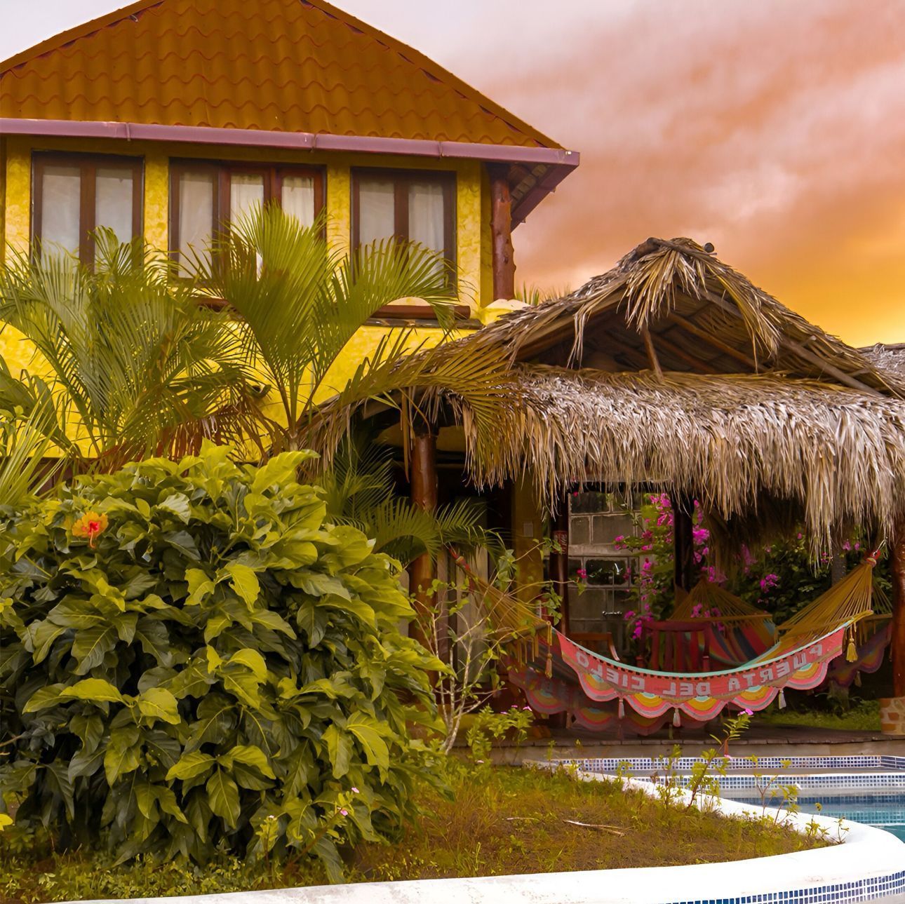 Tropical house with a thatched hut, palm trees, and a colorful hammock at sunset
