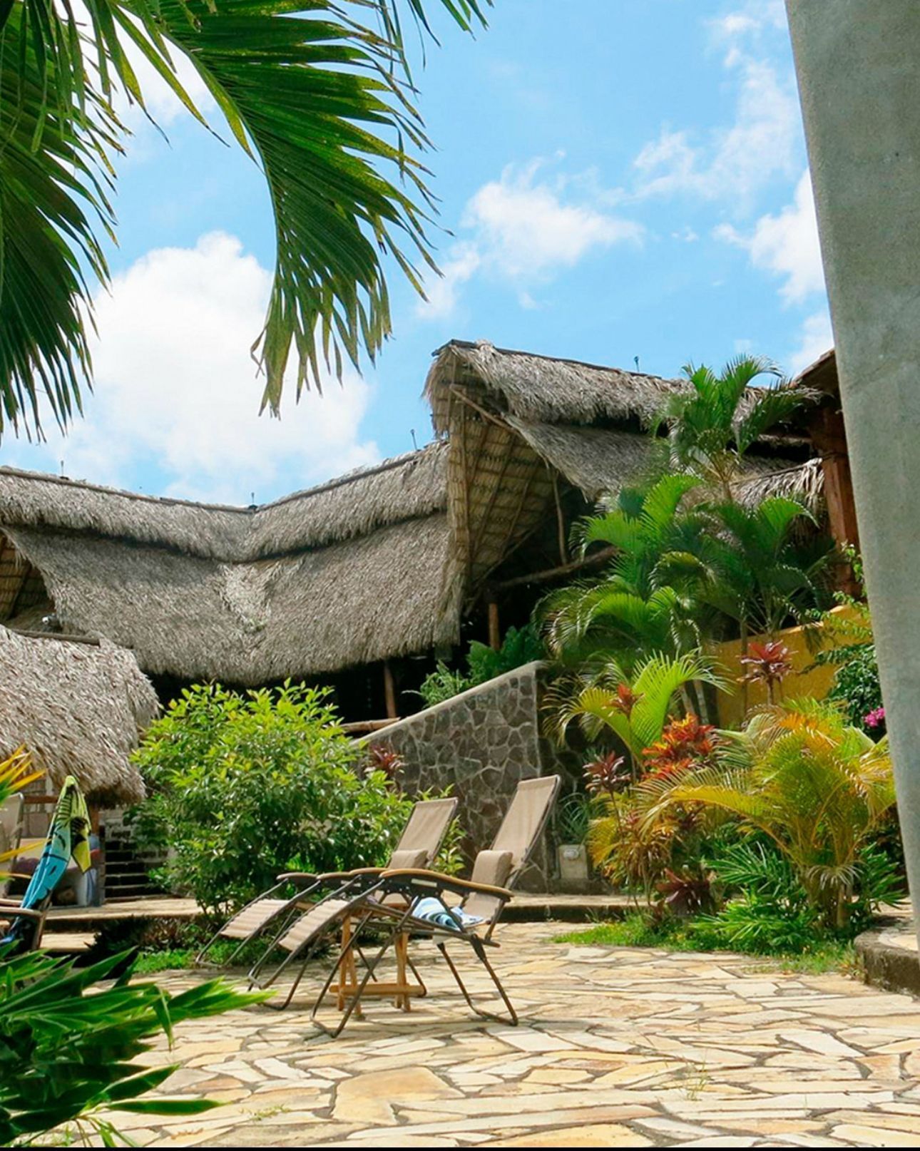 Tropical resort patio with lounge chairs, thatched roofs, and lush greenery under a bright blue sky