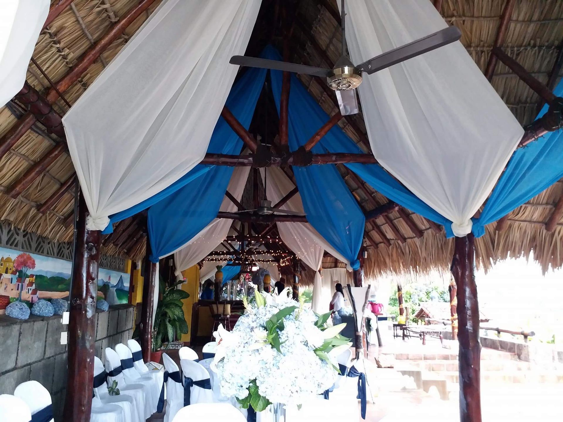 Decorated thatched hall with blue-and-white drapes, banquet tables, and floral centerpieces
