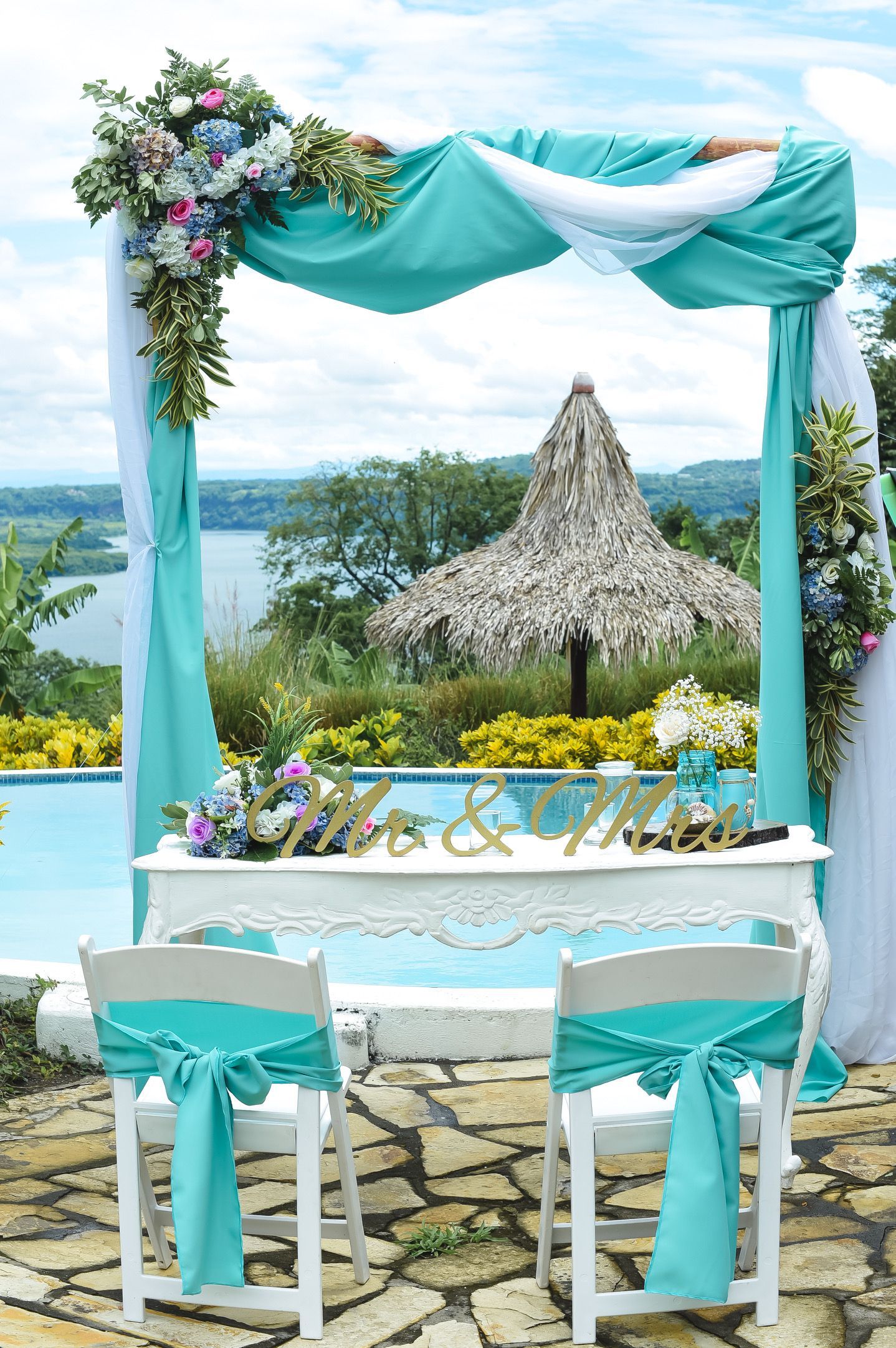 Turquoise wedding arch and table set on a seaside terrace with white chairs and floral decor