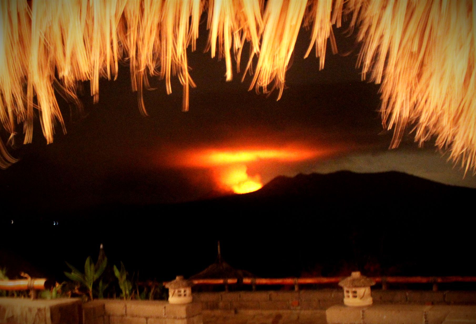 Glowing volcano erupting at night behind dark hills, framed by thatched roof and lit rooftops