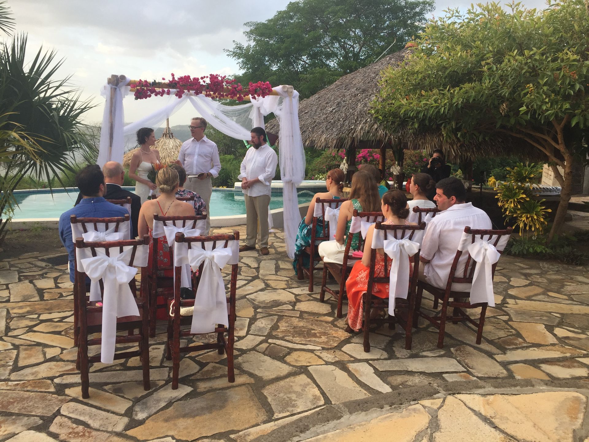 Outdoor wedding ceremony under a floral arch on a stone patio, with seated guests facing the couple.