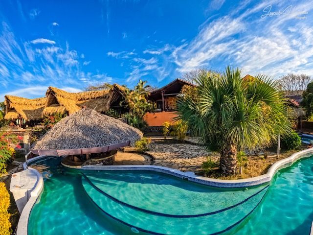 Tropical resort pool with turquoise water, palm trees, thatched huts, and a blue sky