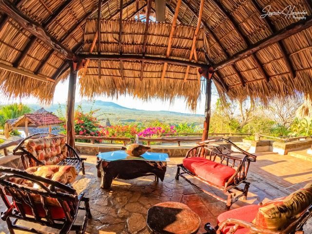 Thatched-roof gazebo with wicker seating and red cushions overlooking a sunny tropical landscape
