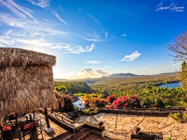 Tropical hillside patio with thatched hut, stone terrace, flowers, and a lake under a bright blue sky