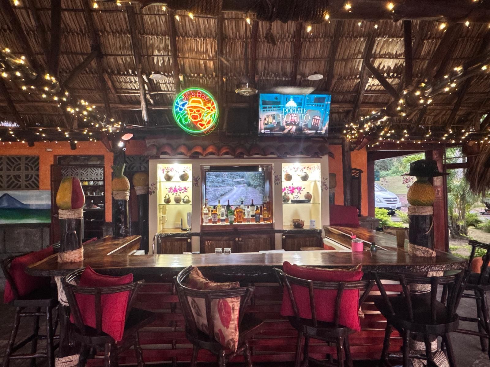 Open-air rustic bar with red chairs, string lights, and TV screens under a thatched roof.