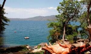 People lounging by a tropical lake with blue water, a small boat, and trees in the foreground.