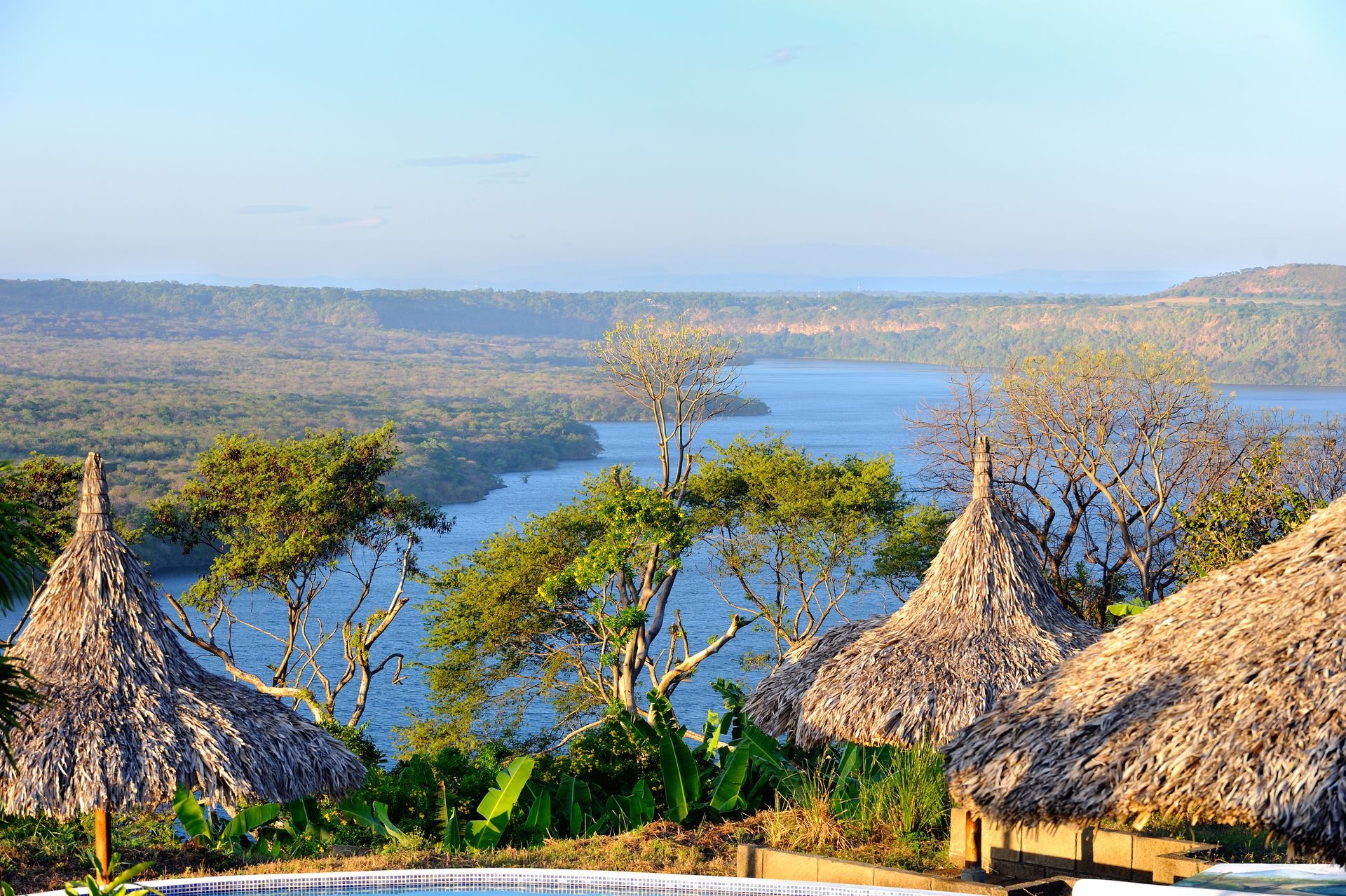 Thatched huts overlooking a blue river valley with trees and hills under a clear sky