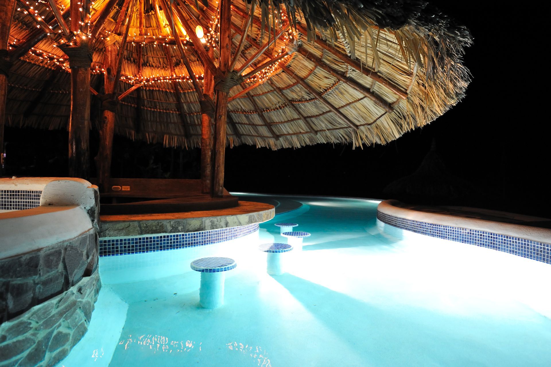 Lit pool with thatched roof hut and palm trees at night