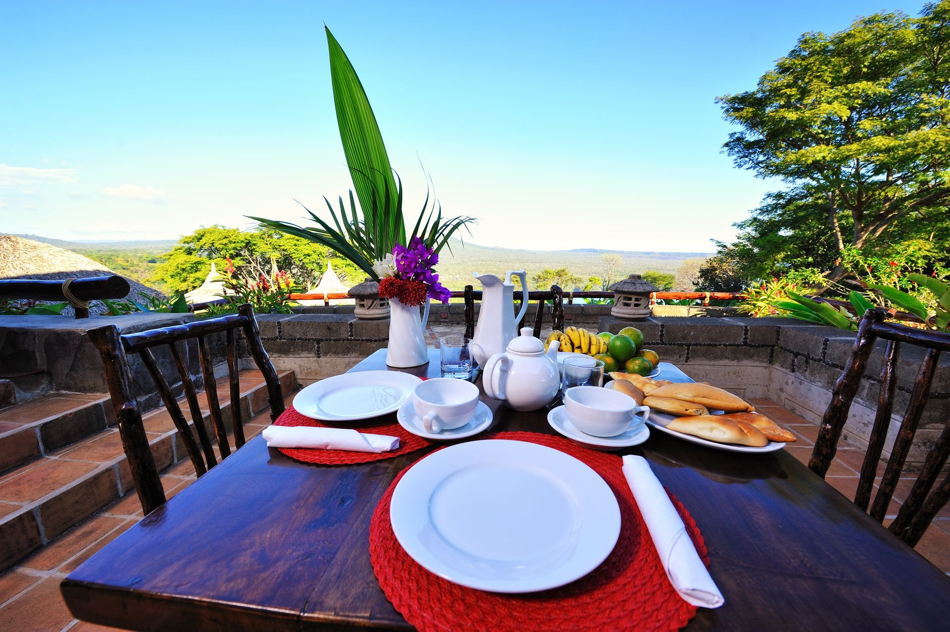 Outdoor dining table set with plates and coffee overlooking a tropical garden and clear blue sky