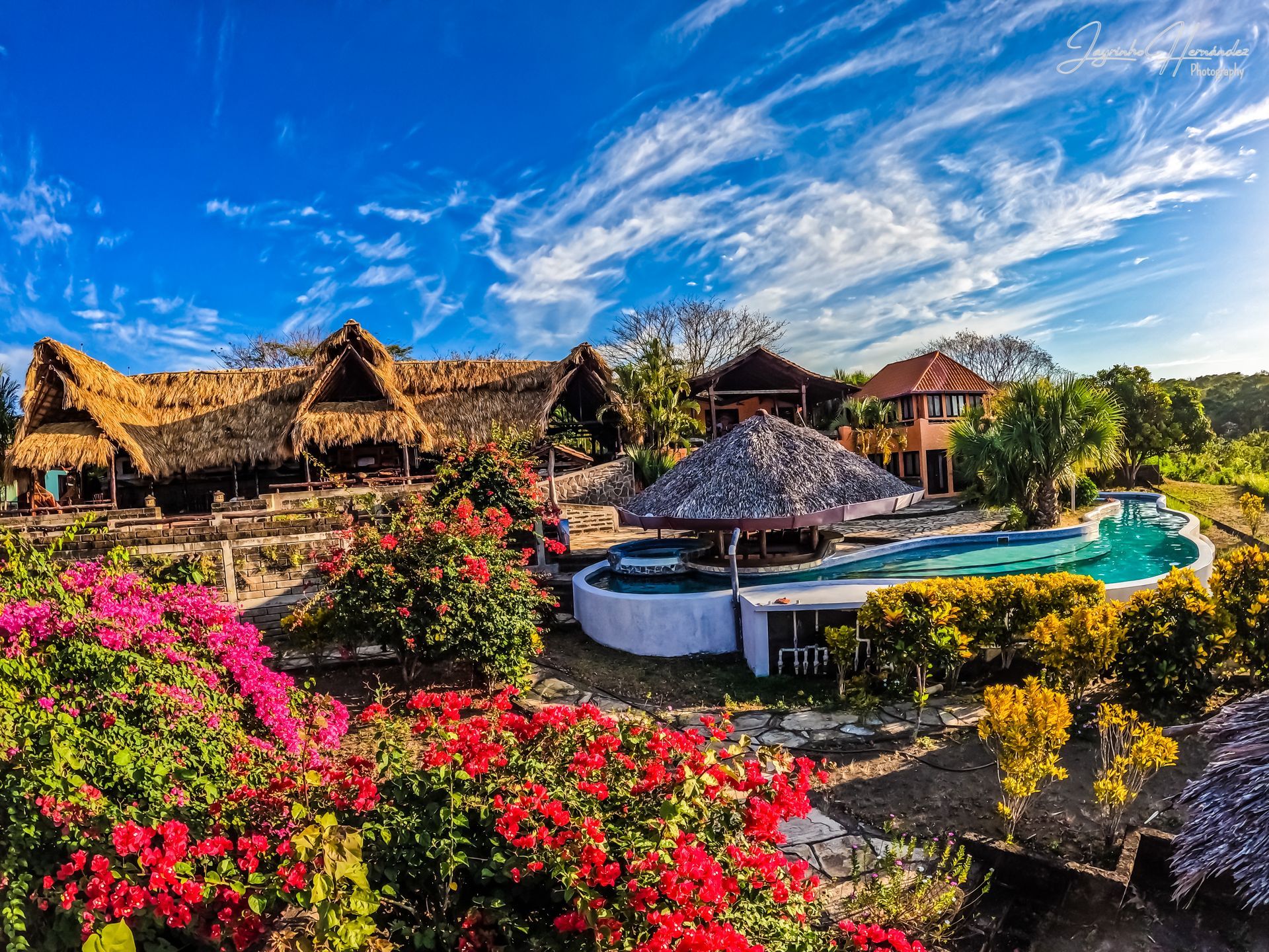 Tropical resort with thatched huts, bright flowers, and a turquoise pool under a blue sky