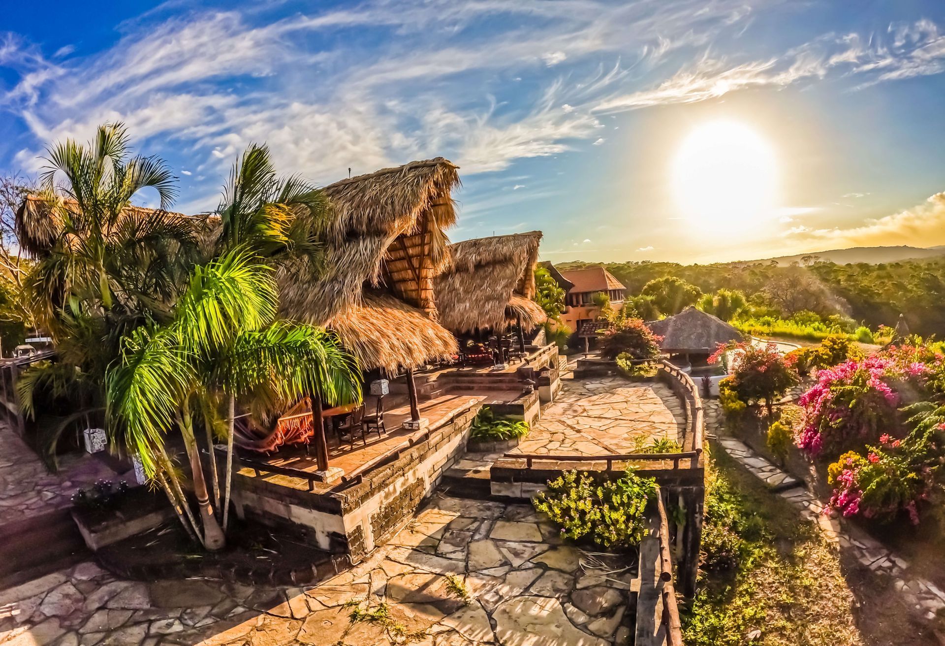 Tropical resort with thatched huts, stone paths, palm trees, and a bright sunset sky.