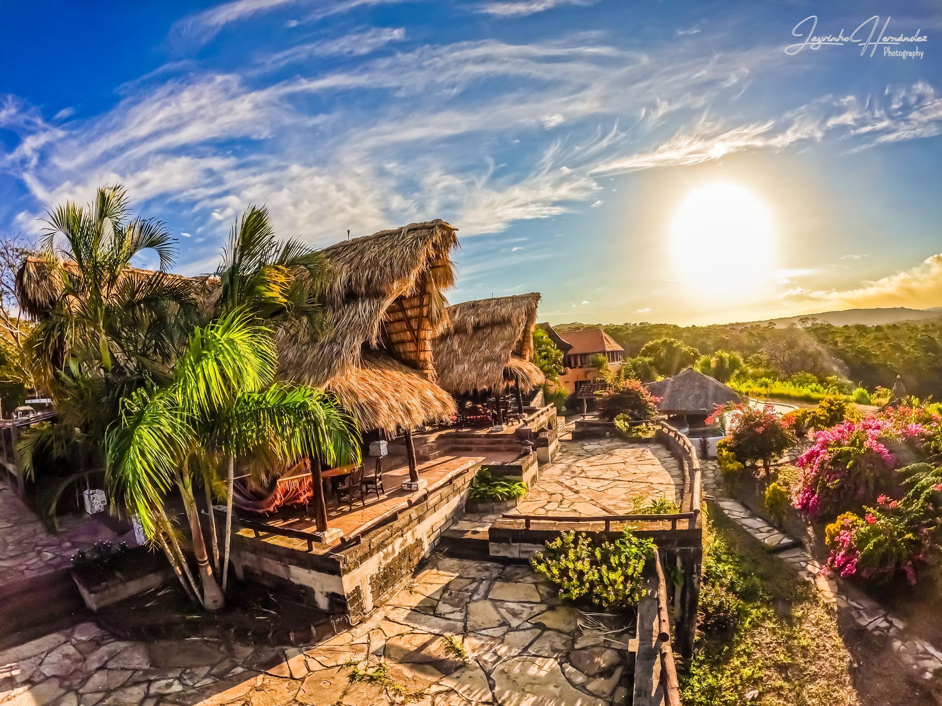 Sunlit tropical resort patio with thatched huts, stone walkway, palm trees, and flowers at sunset