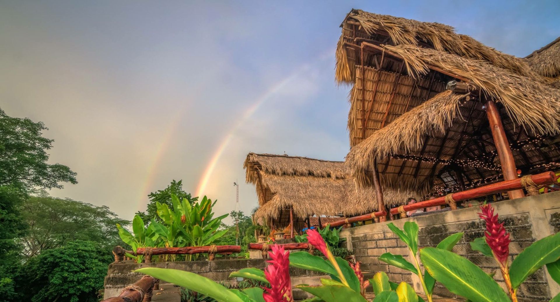 Thatched huts and red flowers in a lush tropical garden beneath a rainbow at sunset