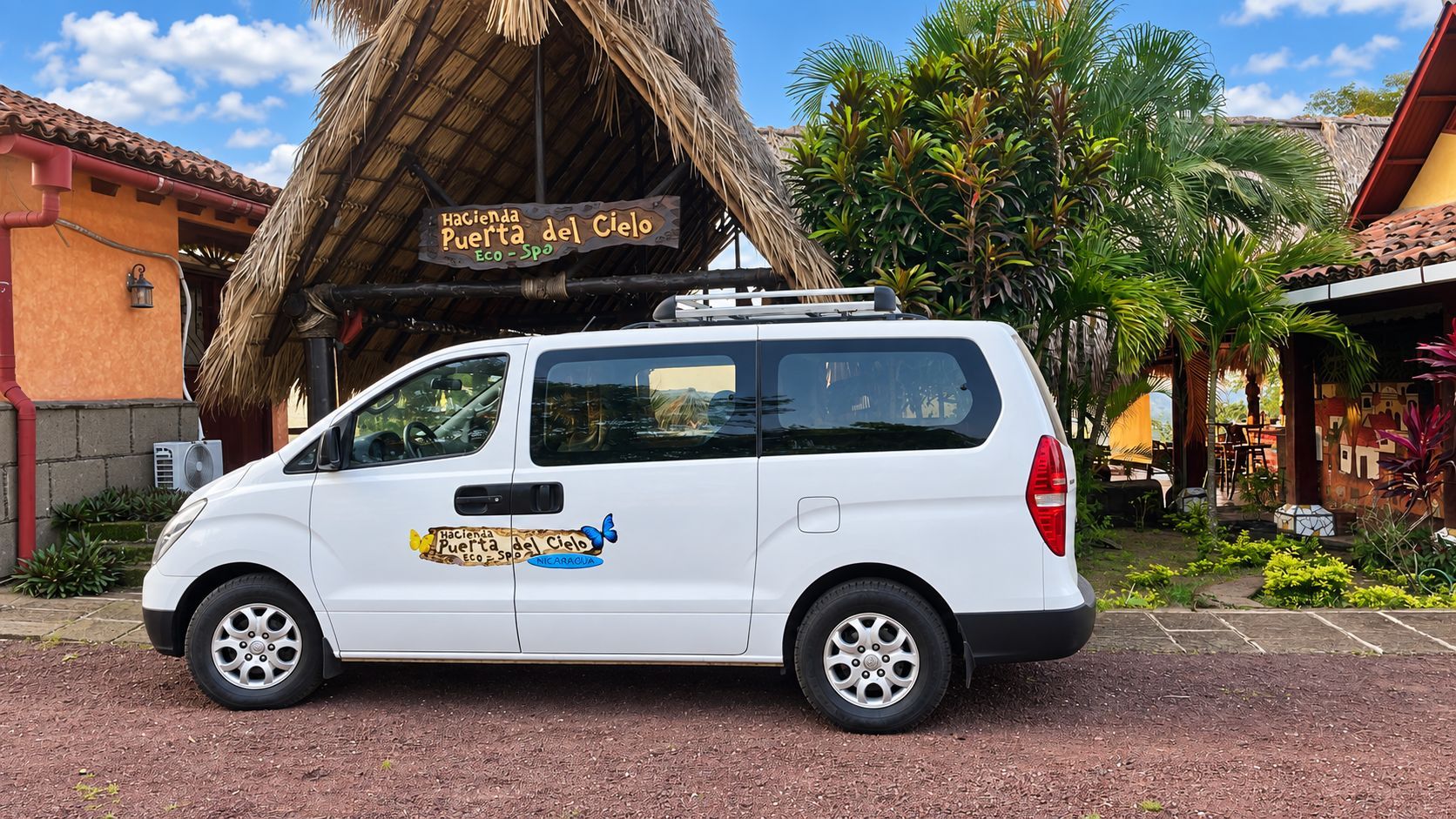 White minivan parked in front of a thatched-roof building with tropical plants and blue sky.