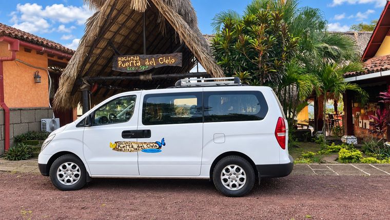 White minivan parked in front of a thatched-roof building with tropical plants and blue sky.