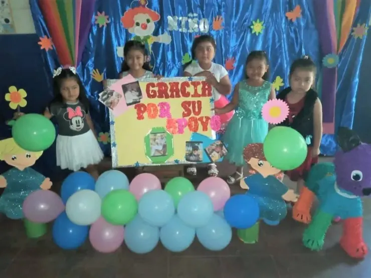 Five children stand on a decorated stage with balloons and a colorful “Gracias por su apoyo” sign.