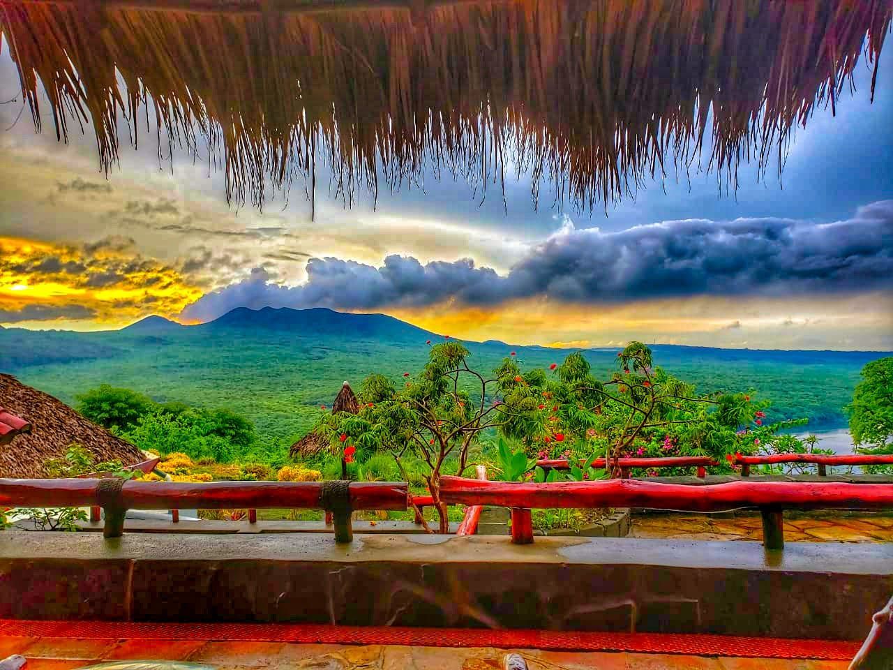 Lush green valley under dramatic clouds, viewed from a thatched terrace with red railing at sunset