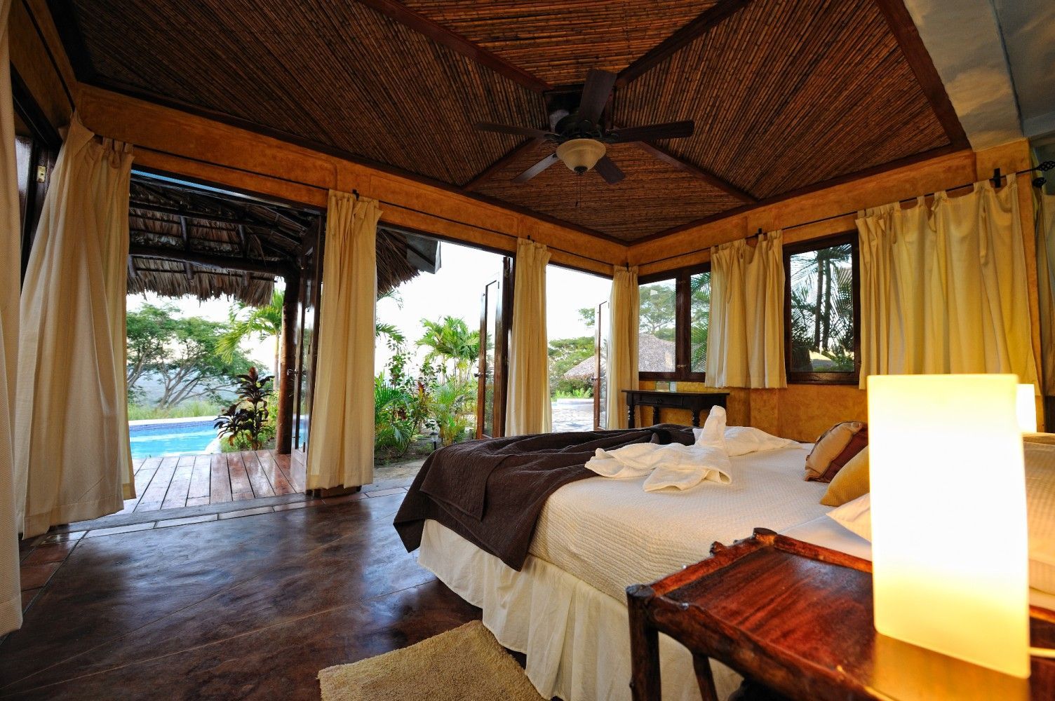 Luxury tropical bedroom with thatched wood ceiling, open curtains, and a view of a pool and palm trees
