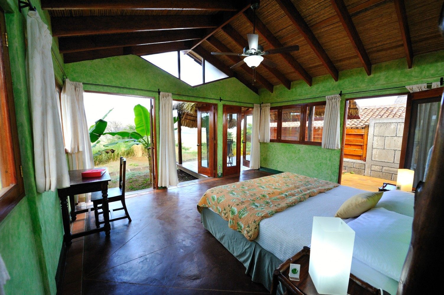Bright bedroom with green walls, wooden ceiling, bed, open doors, and a view of lush greenery outside