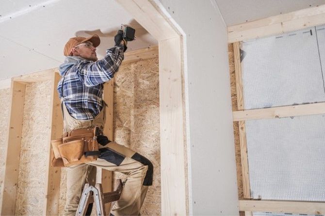 Worker on a ladder installing drywall inside a partially finished room.