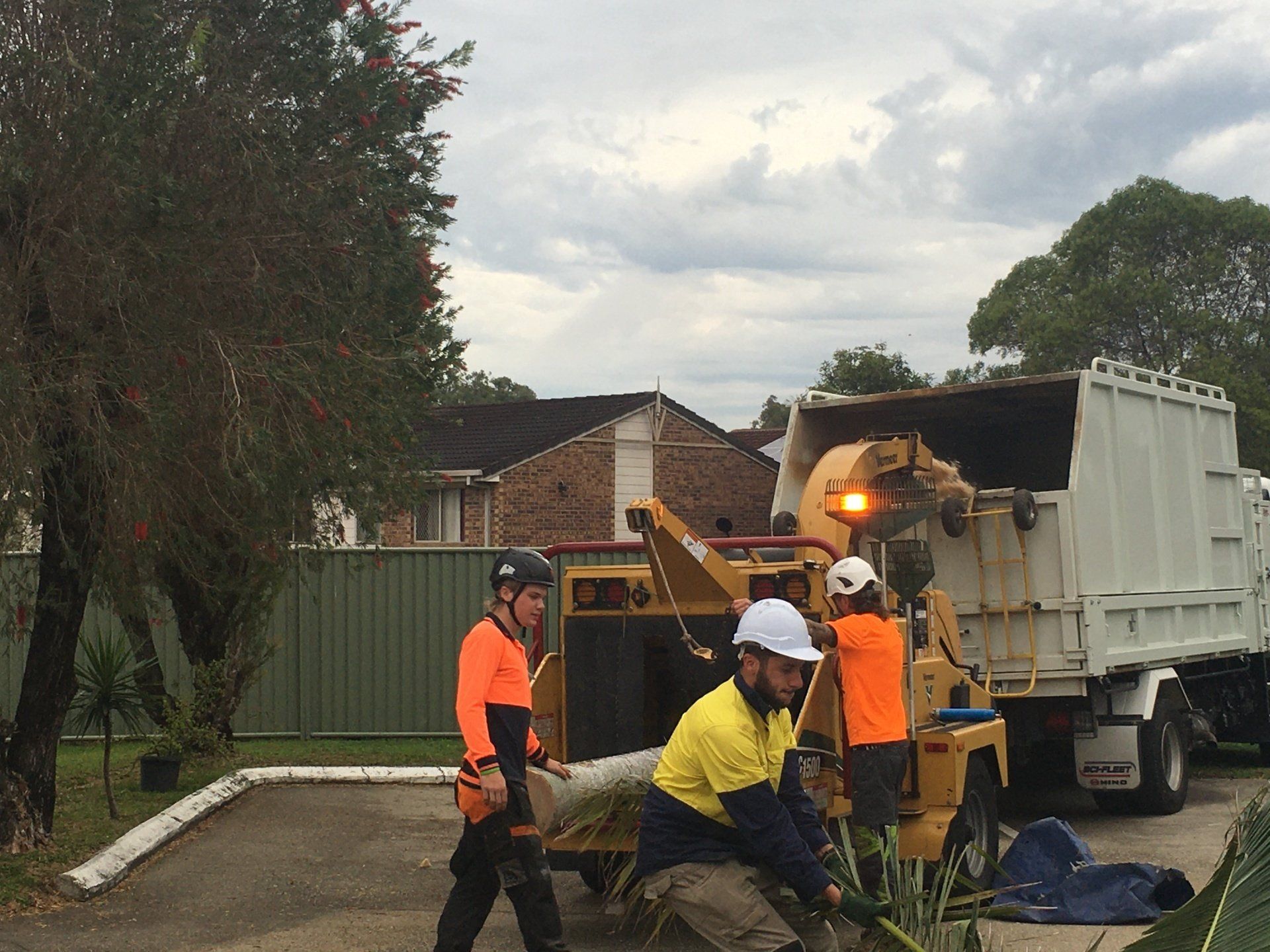 Three workers in high-visibility clothing operate a wood chipper next to a collection truck in a residential driveway.