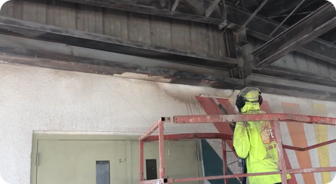 Worker in neon safety gear on a red scaffold inspecting the ceiling of an industrial room.