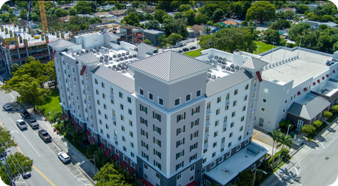 Aerial view of a white multi-story building with a parking lot and trees beside a street.