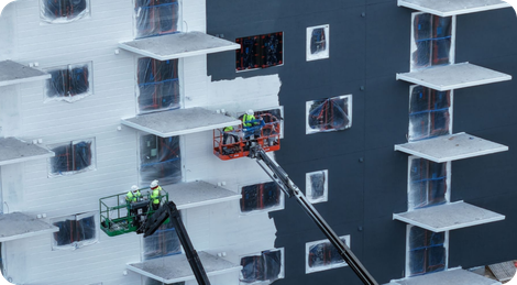Two drones inspect a snowy apartment building facade, one green and one orange, near balconies and windows.
