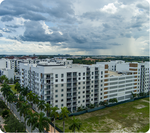 White apartment buildings under a cloudy sky, with palm trees and open grassy land in front.