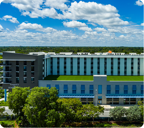 Modern blue-gray building with many windows, green rooftop, and trees under a bright cloudy sky