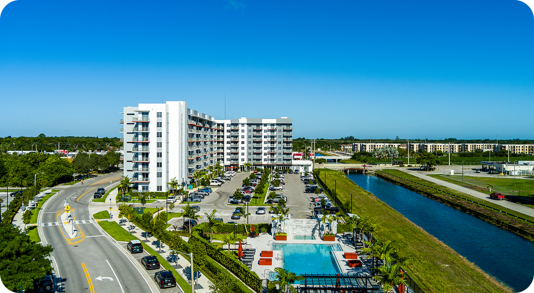 Aerial view of modern white condos, parking lot, and a canal beside a road under a clear blue sky