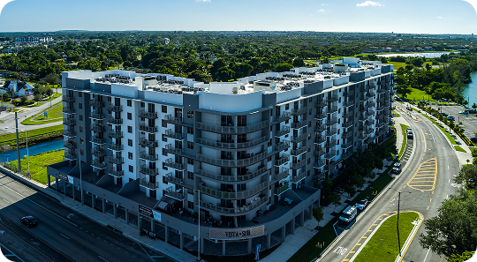 Aerial view of a modern gray apartment building beside a curved road and green landscape.