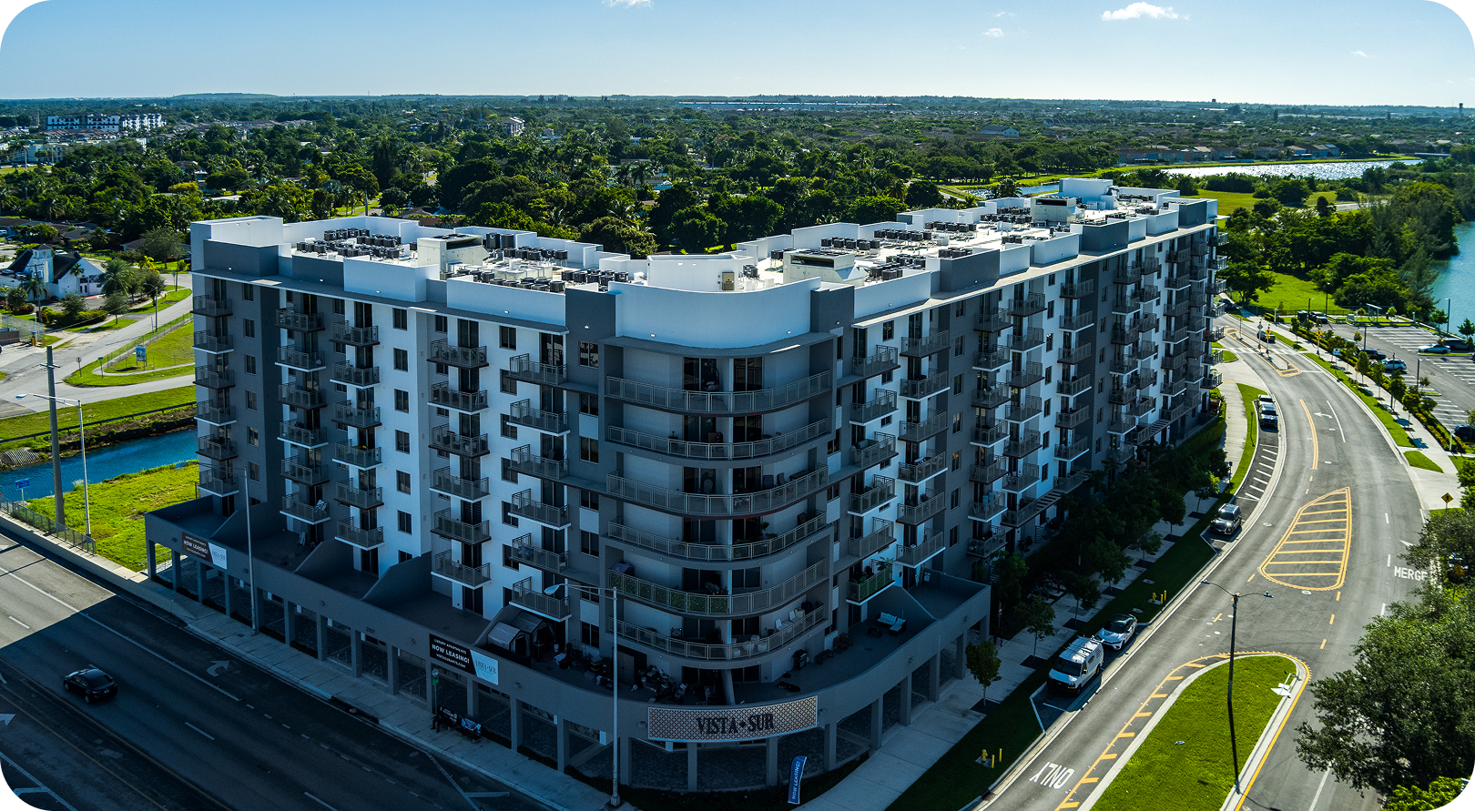 Modern apartment building beside a curved road, surrounded by trees under a clear sky
