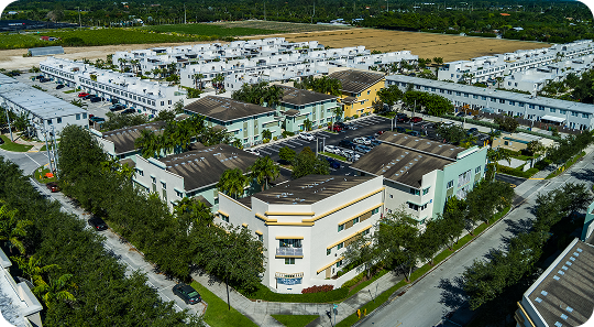 Aerial view of a large apartment complex with white buildings, trees, roads, and green fields nearby.